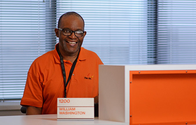 Smiling man wearing an orange The Arc t-shirt sitting at desk behind nameplate that says, "William Washington"