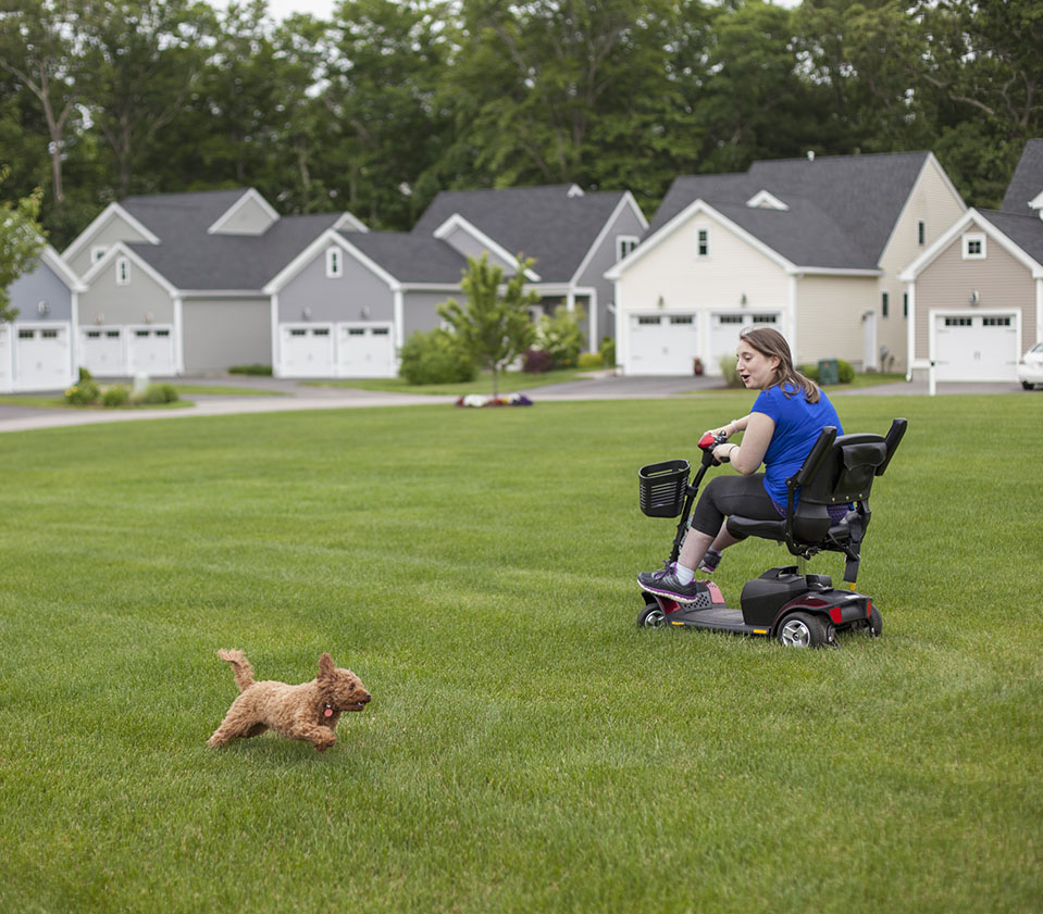 Young woman in scooter playing with small dog on a grassy lawn with houses in the background