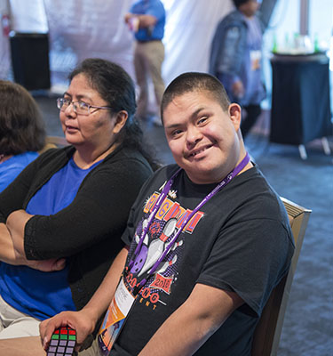 A conference attendee smiles from his seat, wearing a purple lanyard and holding a rubix cube.