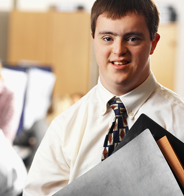 Young man with Down syndrome in business attire smiles at camera holding folders with an office setting in the background