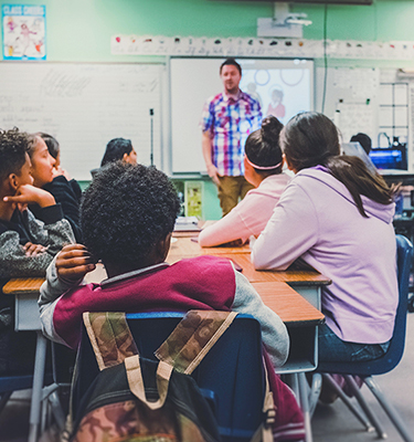 A man stands at the front of a classroom with children sitting at desks listening in the foreground