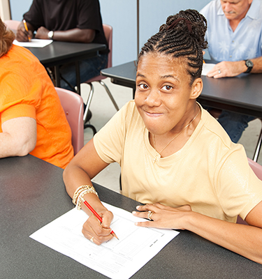 A young woman in a yellow shirt sits at a desk surrounded by other adult students, making eye contact with the camera.