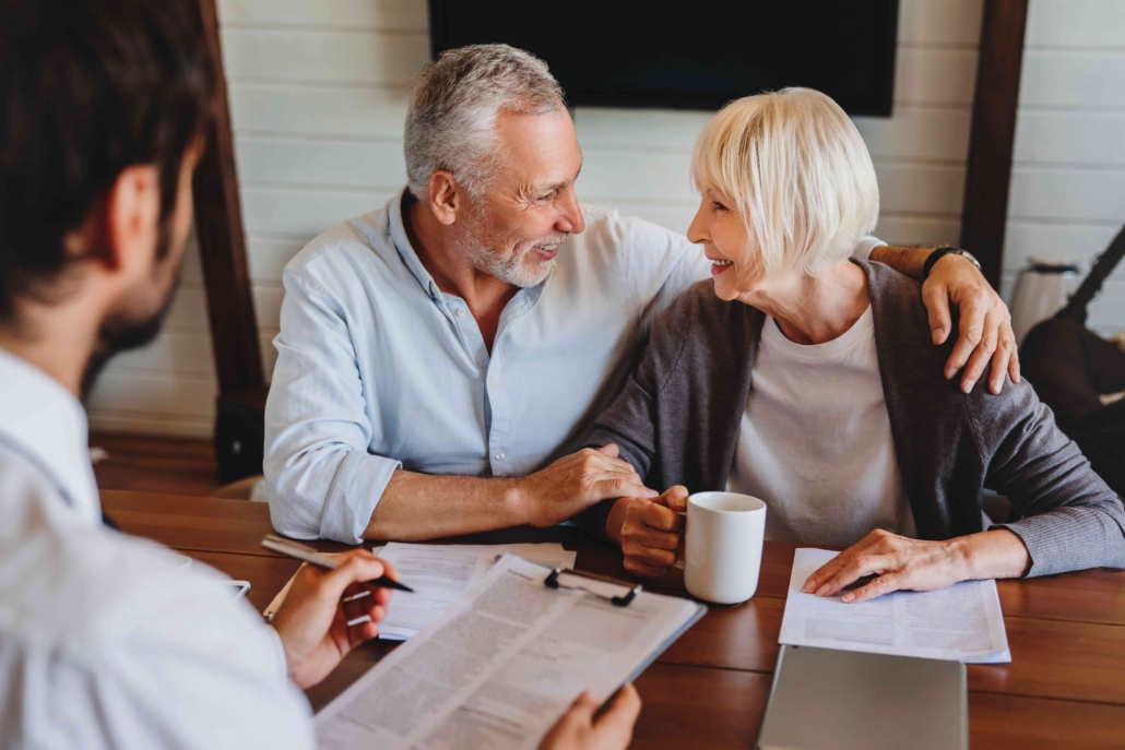 an older couple smiling and looking at each other while sitting at a table with a person across from them holding a clipboard